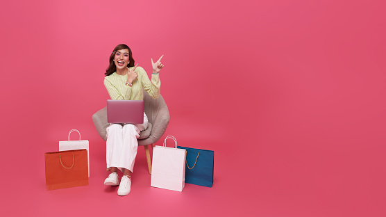 Happy fashionable young asian woman sitting with shopping bag and pointing finger to copy space, she is doing online shopping, Gadget with empty free space for mockup, banner