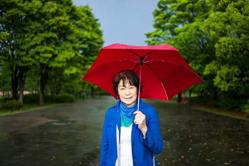Old woman with red umbrella on the street.\nShe is smiling with an umbrella in the middle of the sidewalk in the park.