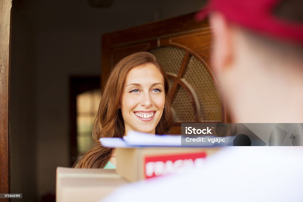 Woman receiving multiple packages at front door Smiling young woman receiving a stack of delivery packages.  The woman has auburn hair and is standing in front of an open residential door.  The male delivery person has short brown hair and is wearing a red visor. 20-24 Years Stock Photo