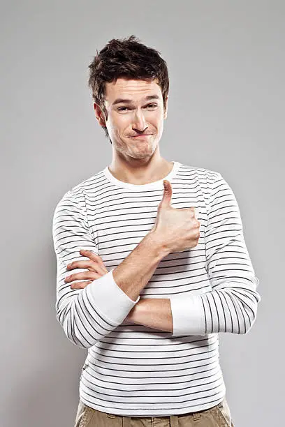 "Portrait of content young man with thumb up, smirking at the camera. Studio shot, grey background."