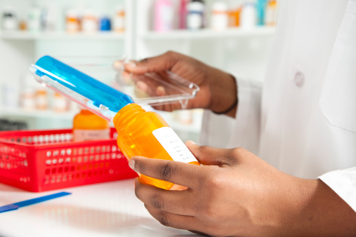 The hands of a pharmacist using a dispensing tool to fill a pill bottle.  The bottle is orange with a printed white label.  It is being filled from a cylindrical blue dispenser with a clear plastic handle.  On the counter is a red plastic basket containing a second pill bottle.