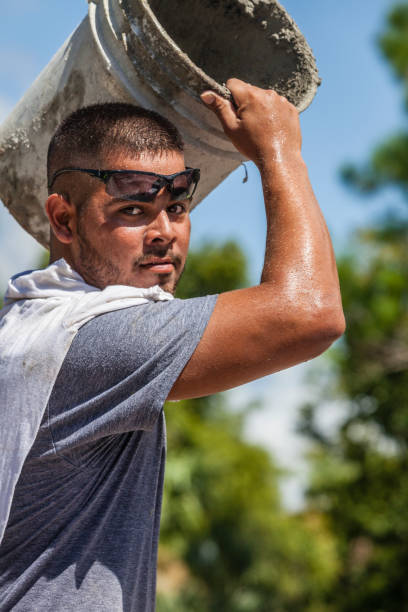 A Hispanic construction worker on a job site in Southwest Florida carries a bucket of cement on his shoulder.