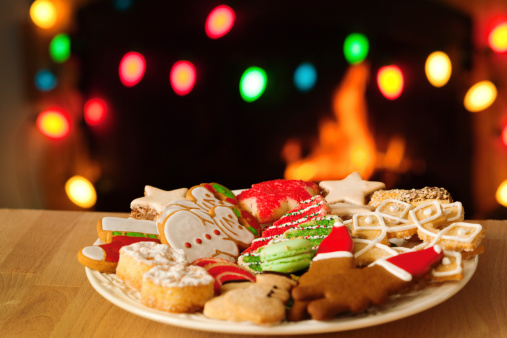 Subject: A tin filled with Christmas sweet cookies decorated with frosting and icing in front of a fireplace decorated with Christmas light.