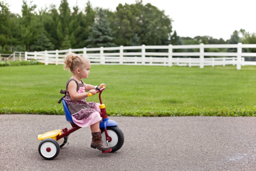 Little girl wearing a dress and cowboy boots riding her tricycle on an overcast morning.