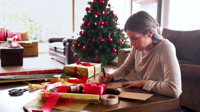 Young woman at home wrapping gifts and writing Christmas cards while sitting on the floor and leaning on a coffee table
