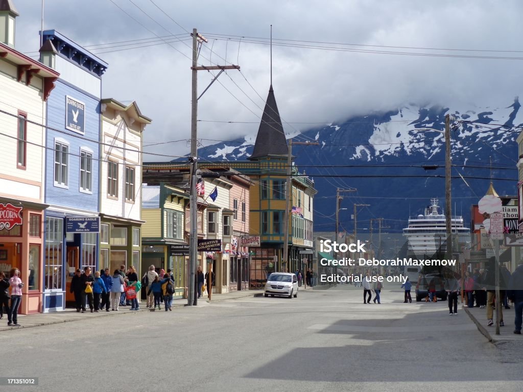 Downtown Skagway Tourists with Snow and Cloud Covered Mountains Background "Skagway, Alaska, United States - July 10, 2012: Streets of downtown Skagway crowded with tourists with a Princess Cruise ship parked in the background in front of snow and cloud covered mountains." Skagway Stock Photo