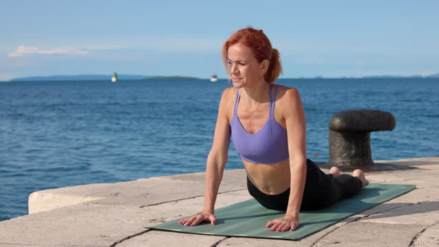 Mature woman lying on yoga mat by the sea in sunshine, lifting upper body and leaning on hands