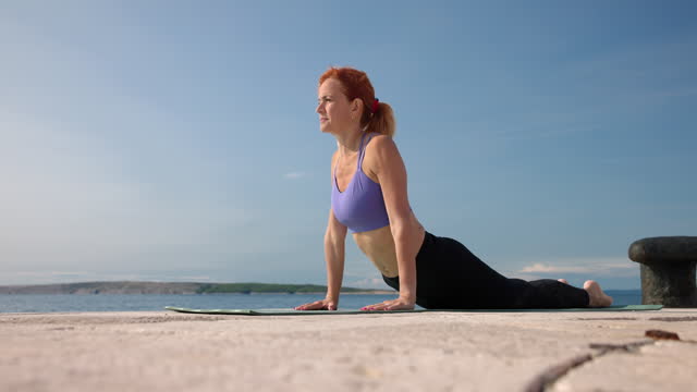 Mid adult woman lying on yoga mat by the sea on sunny day, lifting upper body and leaning on hands