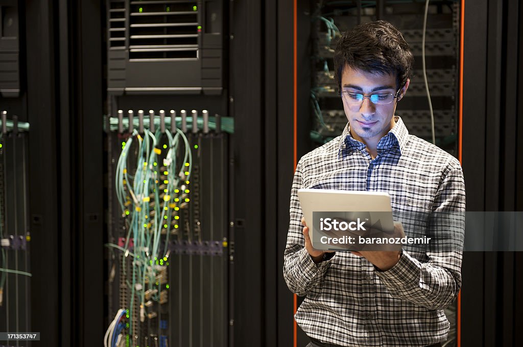 IT Professional Professional IT programming technician is in a server room, checking the main computer equipment.  The man has short brown hair, and he is wearing glasses and a casual black-and-white plaid shirt.  A Digital Tablet is attached to the computer server amid colorful blue computer cables and wires. Network Security Stock Photo