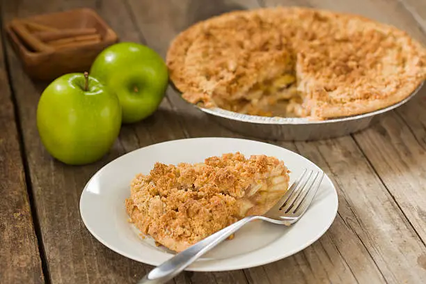 "A high angle close up of a white plate with a slice of freshly baked dutch apple pie, two fresh ripe green granny smith apples, cinnamon sticks and the whole pie in the background, shot on a grungy old wooden table."