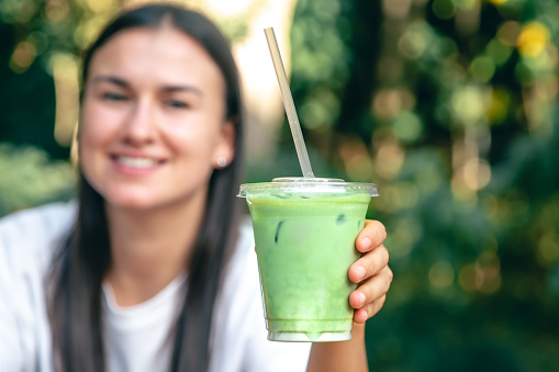 Iced matcha latte in a plastic cup in hands of a woman on a blurred green background, copy space.