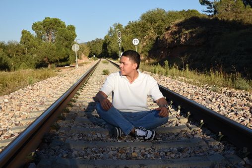 Young young man sitting on the train track on a sunny afternoon, in the mountains