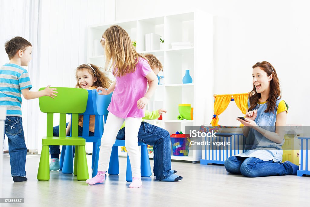 Children Playing Musical Chairs Indoors. Group of preschool children having fun playing Musical Chairs in a preschool, real action, children running. Acceptable blurriness, focus on little girl sitting. Leisure Games Stock Photo