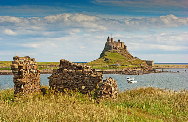 Lindisfarne Castle A view of Lindisfarne Castle on the Holy Isle in Northumberland, England against a pale summer sky with headland and boats in the foreground. northumberland stock pictures, royalty-free photos & images
