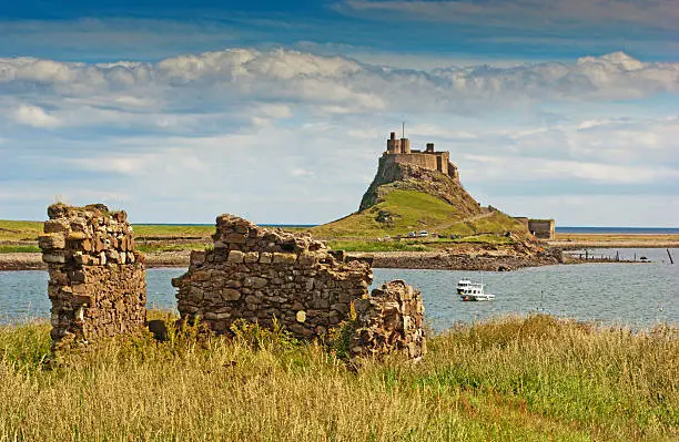 A view of Lindisfarne Castle on the Holy Isle in Northumberland, England against a pale summer sky with headland and boats in the foreground.