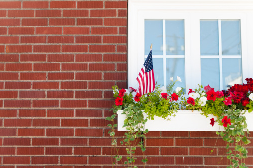White washed window on a brick building with a flower filled window box and american flag sticking out of it.