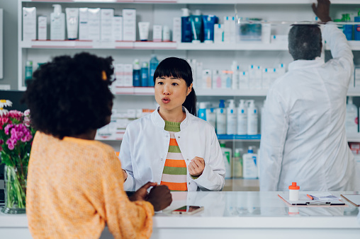Japanese asian woman pharmacist assisting an african american woman customer while standing at the counter in a pharmacy and her male colleague is working near the shelves in the background. Diversity