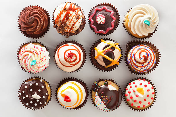 Overhead view of tray with cupcakes Subject: Horizontal high angle, directly overhead view of a dozen frosted cupcakes. The cupcakes are against a white background and are decorated with swirls, sprinkles, drizzles and candies—a tempting decadent treat for those who indulge in sweets and love cakes with lots of toppings. caramel photos stock pictures, royalty-free photos & images