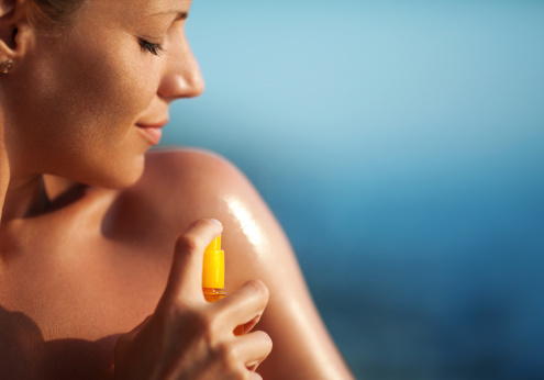 Closeup of adult caucasian woman spraying suntan lotion onto her shoulder. It's sunny summer day and she's sunbathing with a smirk on her face. Blurry sea in background.