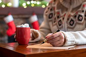 Woman writing letter to santa claus using fountain pen on sheet of paper at christmas fireplace with decoration of light bulbs drinking hot cocoa and marshmallow.