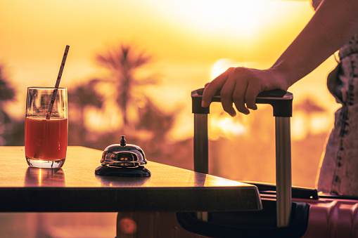 Woman with suitcase ringing hotel service bell with sea and palm tree view on sunset. Travel concept. 24-hour beach hotel front desk. Late check-out.