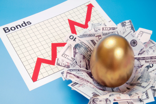 On a light blue background rests a golden egg in a nest made of money which is beside a white piece of paper outlining the stock market graph which is shown in red to be moving up and down