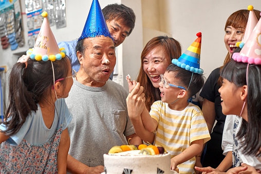Multi-generation Asia family hold a birthday party to celebrate a boy’s birthday at home. A little boy putting cake cream on grandfather face and nose on his birthday party. They are wearing birthday hats and having fun together. It’s a happy theme about love concept, emotion, togetherness, life, enjoyment, Affectionate. There is father, mother, son, daughter, grandfather in this photo. There is a birthday cake on the table. The girl twins are indigenous in Taiwan.