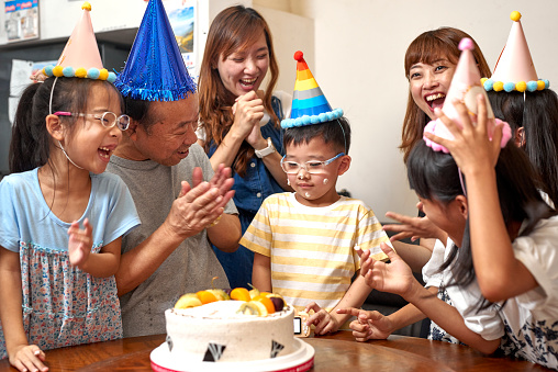 Multi-generation Asia family hold a birthday party to celebrate a boy’s birthday at home. A grandfather putting cake cream on a little boy face and nose. They are wearing birthday hats and having fun together. It’s a happy theme about love concept, emotion, togetherness, life, enjoyment, Affectionate. There is father, mother, son, daughter, grandfather in this photo. There is a birthday cake on the table. The girl twins are indigenous in Taiwan.