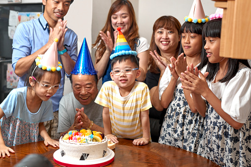 Multi-generation Asia family hold a birthday party to celebrate a boy’s birthday at home. The mother and boy make a wish on his birthday party. They are wearing birthday hats and having fun together. It’s a happy theme about love concept, emotion, togetherness, life, enjoyment, Affectionate. There is father, mother, son, daughter, grandfather in this photo. There is a birthday cake on the table. The girl twins are indigenous in Taiwan.