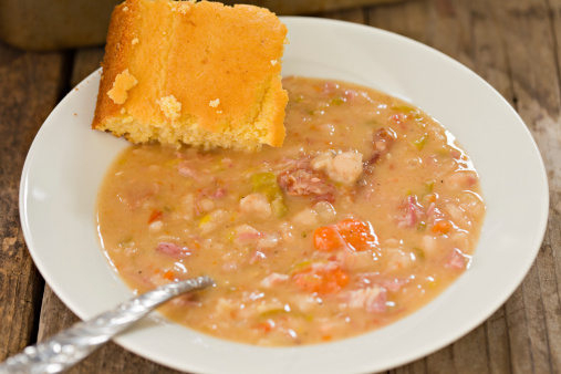 A high angle close up shot of a white bowl of freshly made navy bean soup and a fresh slice of golden brown corn bread.