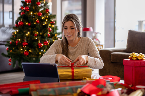 Happy Latin American woman at home wrapping Christmas gifts following an online tutorial