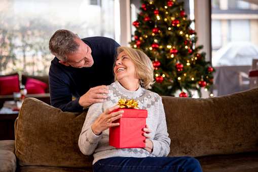 Mature couple celebrating Christmas and man surprising his wife with a present - holiday season concepts
