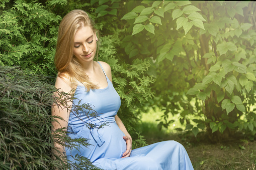 Pregnant young woman dressed in a blue dress is sitting in the garden.  Horizontally.