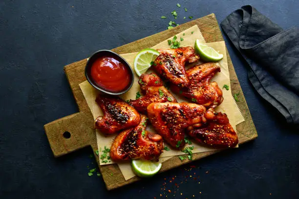 Grilled spicy chicken wings in mexican or chinese stile with ketchup on a wooden cutting board on a dark slate, stone or concrete background. Top view with copy space.
