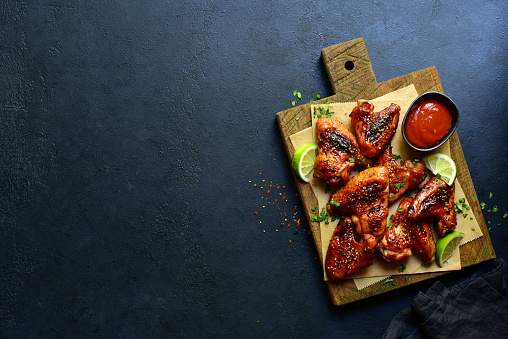 Grilled spicy chicken wings in mexican or chinese stile with ketchup on a wooden cutting board on a dark slate, stone or concrete background. Top view with copy space.