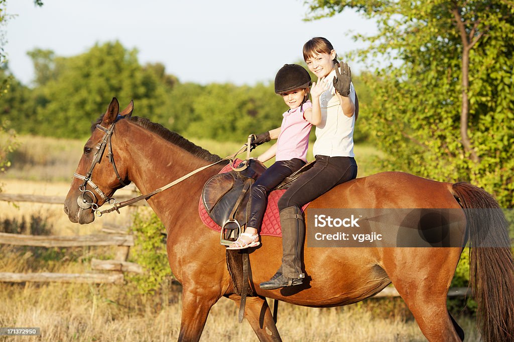 Child Riding Horse Outdoors. Little girl with riding hat learning how to ride a horse. Riding horse together with her instructor. Little girl and instructor looking at camera and waving. A Helping Hand Stock Photo