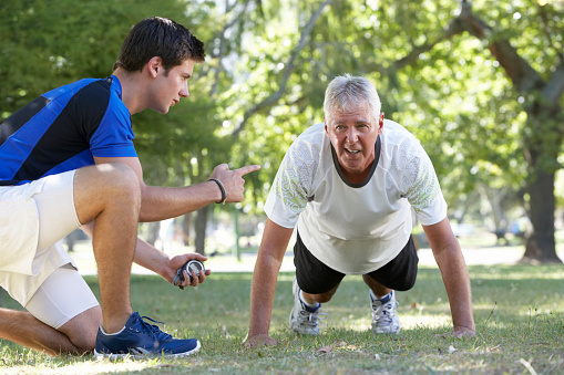 Senior Man Working With Personal Trainer In Park Doing Press Ups