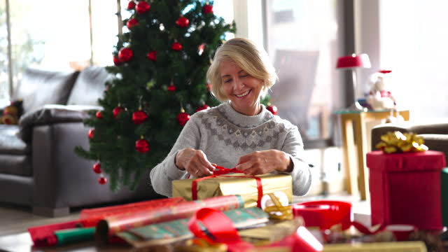 Happy senior woman at home sitting on the ground packing Christmas gifts smiling very cheerfully full of the Christmas spirit