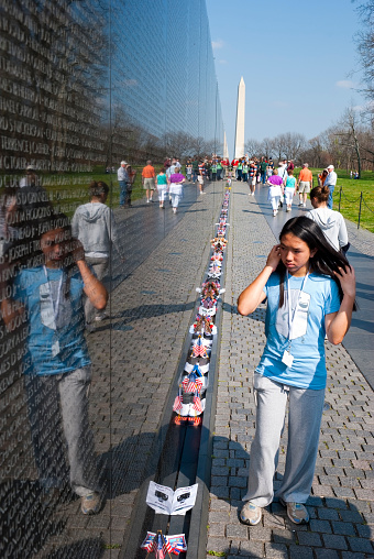 Washington DC, USA - April 10, 2008: A young girl of Asian descent walks along the Vietnam Veterans Memorial, her reflection visible in the wall inscribed with names of the dead. Other visitors and the Washington Monument are in the distance.