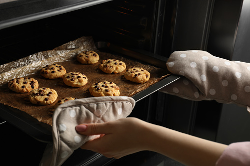 Woman taking out delicious chocolate chip cookies from oven, closeup