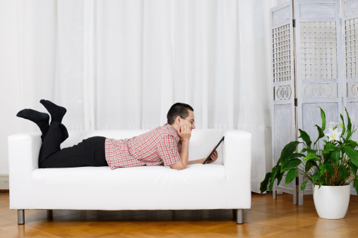 Black-haired Man laying on White Sofa looking to Tablet PC, Germany