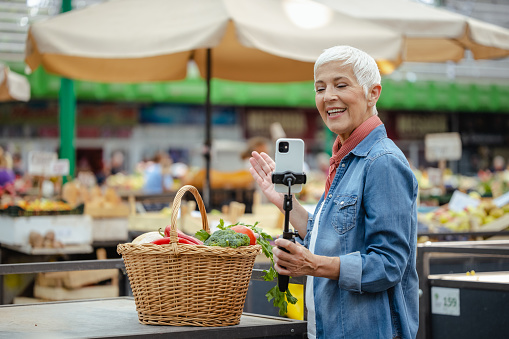 A cheerful senior Caucasian female vlogger filming herself standing at a local marketplace. She is holding a selfie stick and standing next to a woven basket filled with vegetables.