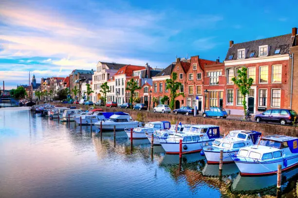 "Historic cityscape along a channel in Delfshaven, a district of Rotterdam, the Netherlands. Visible are typical dutch architecture, historic sailing boats,windmill, restaurants, colorful reflection in the river, blue and dramatic cloudscape and beautiful sunset atmosphere."