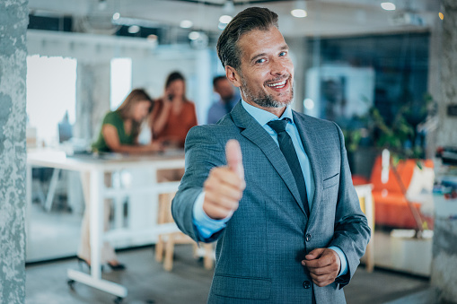 Portrait of a young businessman in the modern office, and a team working behind him. Successful businessman standing in creative office smiling with thumbs up and looking at camera.