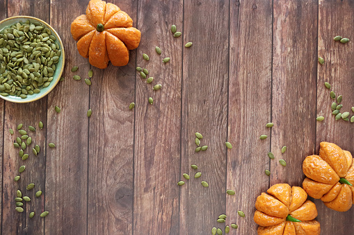 Stock photo showing close-up, elevated view of border of freshly baked, homemade bread pumpkin designed rolls with green chilli pepper stalks and pumpkin seeds made into a poster frame, against a woodgrain background.