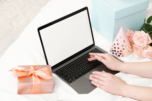 Woman using laptop near gift and rose flowers on bed in room, closeup. Happy Birthday