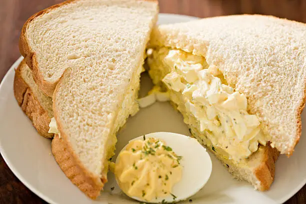 A high angle extreme close up shot of an egg salad sandwich on white bread and a deviled egg.