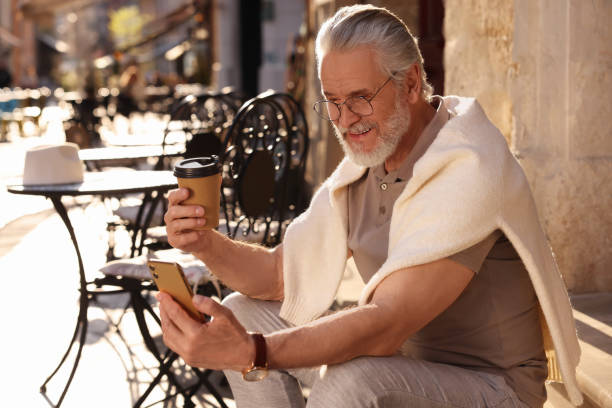 istock Handsome senior man sitting on doorstep, using smartphone and drinking coffee outdoors, space for text 1713847654