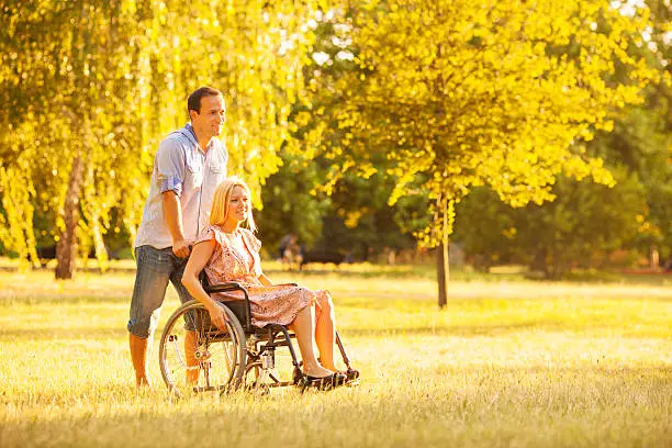 Portrait of an young couple, man and disabled woman in wheelchair walking in a park.