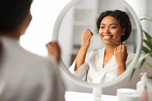 Oral Care. Smiling Black Woman Using Dental Floss Near Mirror At Home, Beautiful Young African American Female Cleansing Teeth, Making Daily Hygiene Routine, Seletive Focus On Reflection